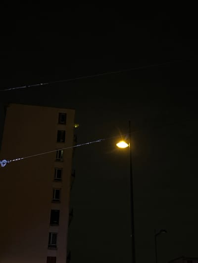 A nighttime shot shows a building with small, regularly spaced windows, illuminated in a warm, orange glow. Above, a bright yellow light hangs from a dark horizontal line. Cables stretch diagonally across the frame, connecting to the light and extending downwards. The background is a deep, unlit darkness.