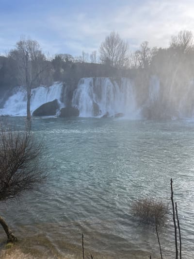 The image depicts Kravica Waterfall in Zvirići, Bosnia and Herzegovina, viewed from the river level. A wide, multi-tiered waterfall cascades into a turquoise-colored river that dominates the mid-ground and foreground. Prominent water mist and spray rise from the powerful falls, particularly on the right side, partially obscuring the distant trees.
The background features a dense line of bare deciduous trees along the top of the waterfall and its surrounding banks, indicating a cold season. A tall, bare tree stands distinctly on the left bank, partially submerged in the water near the falls. The sky above is light blue with sparse cloud cover, suggesting a bright day, possibly with low sun causing some atmospheric haze or glare on the right.
In the foreground, the river water shows ripples and reflects light. The shallow left bank reveals submerged sandy or muddy ground. Smaller, bare shrubs and individual branches protrude from the water and riverbank in the immediate foreground, anchoring the scene. No individuals or man-made structures are present within the frame.