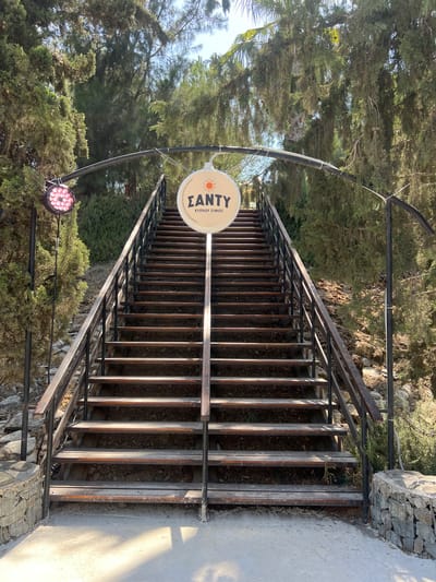 An exterior shot captures a set of wooden stairs with metal railings leading upwards, flanked by trees and greenery. A circular sign hangs above the stairs, featuring a sun graphic and Greek text. To the left of the stairs, a large, circular light fixture with pink LEDs is mounted on a pole. The scene appears to be located outdoors, possibly in a park or at an establishment. The text on the sign reads "ΣΑΝΤΥ" and below that, "ΚΥΠΡΟΥ ΣΥΘΟΣ". This sign likely indicates the name of a place or event. The overall impression is that of an entrance or pathway to a venue, set against a natural, sun-dappled background.