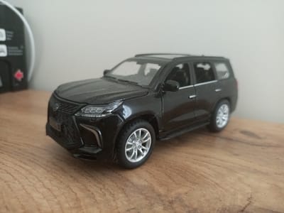 A black toy SUV, identified as a Lexus LX 570 by the license plate, is positioned on a wooden surface. The toy car is black with silver wheels and detailed headlights and grille. In the background, a light gray wall is visible, and to the left, a partially seen white circular object with black markings is present. The scene appears to be indoors, with natural lighting. There is no indication of this image being associated with Usolye-Sibirskoye, Russia.