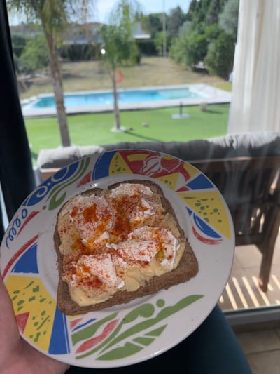 The image shows a piece of whole-wheat toast on a white plate with a colorful pattern. The toast is spread with a yellow substance, possibly hummus or a similar spread, and topped with dollops of white cheese or cream cheese, sprinkled with red paprika. The plate is held by a person. Through a window, a blurred view of an outdoor area is visible, including a swimming pool, green grass, and trees, suggesting a residential setting. The location is Urbanización Encinares De Alcolea, Spain.