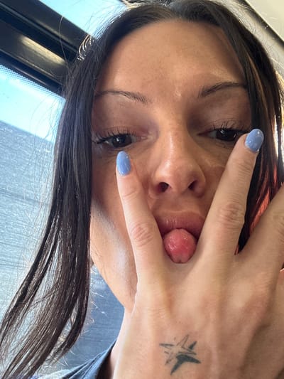 An close-up, angled portrait captures a person, likely female, with dark hair cascading around their face. The subject is seen from a slightly upward angle, with their head tilted. Their left hand partially covers their face, with the index and middle fingers outstretched, forming a "peace" sign or similar gesture, and the thumb and pinky finger obscuring the lower part of their face. Their tongue is sticking out playfully between their fingers.
The person has dark eyes with prominent eyelashes and well-defined eyebrows. Their skin tone appears warm, and there's a visible sheen on their forehead and cheek, possibly from natural oils or lighting. The visible fingernail on the index finger is painted with a light periwinkle or baby blue polish. On the back of the hand, near the wrist area, a small, faded black tattoo resembling an abstract star or burst pattern is visible.
The background is largely out of focus but shows bright daylight coming through what appears to be a window. A dark window frame is visible on the left side, and reflections of the sky or light sources are present. A hint of a dark blue or grey garment can be seen on the right edge of the frame, possibly a shoulder or collar. The overall mood is playful and informal. There is no visible text in the image. Given the provided context, the image was taken in Sevenoaks, United Kingdom, during daytime.