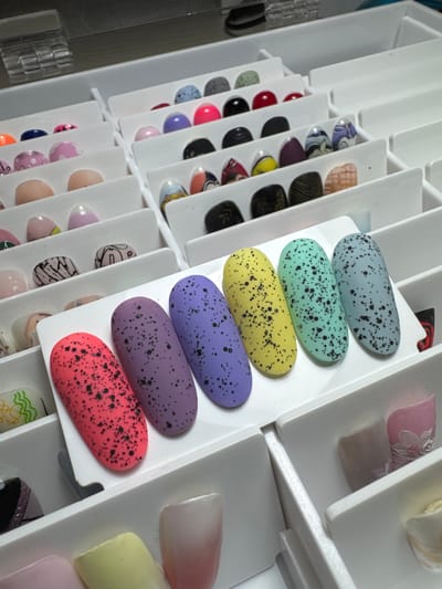 The image displays a collection of artificial fingernails organized in white compartments, likely a nail art display or storage unit. The fingernails are varied in color and design, featuring solid colors, speckled patterns resembling Easter eggs, and intricate artistic designs. In the foreground, four speckled nails are prominent: bright pink, purple, yellow, and light green. To the right of these, a light blue speckled nail is visible. In the background, other compartments hold more fingernails with different styles. The environment appears to be indoors, with artificial lighting. There is no indication of Vlasikha, Russia, or any specific city. The image is a close-up shot focusing on the fingernails.