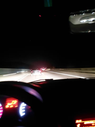 A nighttime view from inside a vehicle shows a highway through the windshield and the dashboard of the car. The outside scene reveals other cars with their headlights and taillights visible on the road. The dashboard features illuminated gauges and digital displays. A rearview mirror is partially visible on the left side. The steering wheel is also visible.