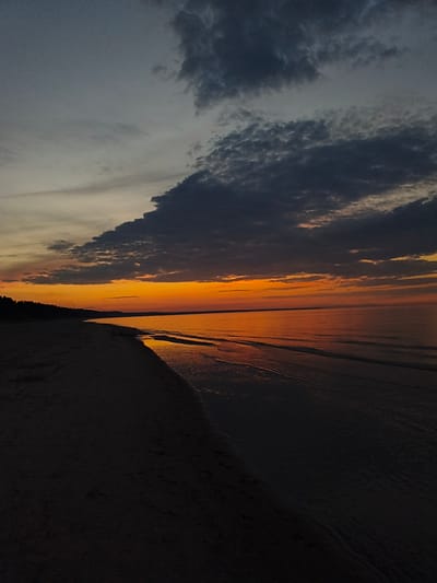 The image depicts a beach at dusk in Apšuciems, Latvia. The sky is a gradient of colors, transitioning from dark grey clouds at the top to a vibrant orange sunset near the horizon. A dark silhouette of trees lines the left side of the image, marking the edge of the land. The beach itself is dark, with the wet sand near the water reflecting the sunset's glow. Small waves gently lap the shore, creating a calm and serene atmosphere. There are no people or specific objects visible in the frame.