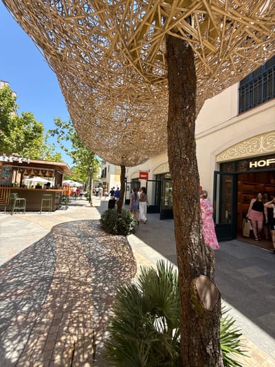 A sun-drenched pedestrian street in La Roca Village, La Roca del Vallès, Spain, is depicted. A large tree trunk dominates the foreground, its bark textured and rough, with a recent cut visible on its side. Above the tree, an elaborate canopy made of woven natural material casts intricate shadows on the cobblestone ground. To the left, a small food stall with green stools and umbrellas is visible, with customers browsing nearby. Further down the street, shoppers walk past storefronts, one of which displays the "HOF" logo. The architecture is characterized by light-colored buildings with dark-shuttered windows and decorative moldings. The sky is a clear, bright blue. A woman in a pink floral dress is partially obscured by the tree trunk, facing away from the camera. Other individuals are seen walking and shopping, engaged in typical street activity. Lush green trees line the sides of the street, adding to the pleasant outdoor ambiance.
