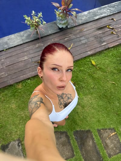 A female individual is depicted in an outdoor setting, likely a private residence or villa, in Kerobokan, Indonesia. She is positioned low, appearing to be kneeling or crouching on green grass, and holds a camera at arm's length, angled upwards for a selfie. Her red hair is pulled back, revealing blue eyes looking directly at the camera, accentuated with eyeliner and mascara. Her lips are slightly parted in a pout, displaying a light-colored lipstick. She wears a small golden ring in her left nostril. Her left ear features multiple gold-toned helix piercings and a prominent golden ear gauge in the lobe. She is dressed in a white, thin-strapped top. Visible tattoos include a large black ink bird design on her left upper arm/shoulder and a large black ink floral design on her chest/sternum.
The background reveals a bright blue swimming pool to the upper left, bordered by a dark grey concrete coping. Adjacent to the pool is a wooden deck constructed from dark planks, upon which several dry leaves are scattered. Two potted plants, one with green and reddish foliage in a clear plastic bottle, and another with lighter green leaves, are situated near the pool's edge. The foreground and right side of the image consist of well-maintained green grass. Several dark grey, rectangular stepping stones are embedded within the grass, receding into the distance. Additional dry leaves are scattered across the grass.