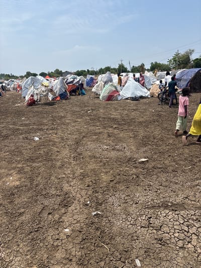The image shows a refugee camp in Al Babali, Sudan, with numerous makeshift tents and shelters. The ground is dry and cracked, indicating arid conditions. Several people, including adults and children, are visible among the tents. Some are seen walking, while others are gathered near the shelters. The tents are made from various materials, including tarps, plastic sheeting, and fabric, and are set up close together. The overall scene depicts a temporary settlement established in a rural or undeveloped area, typical of displacement situations.