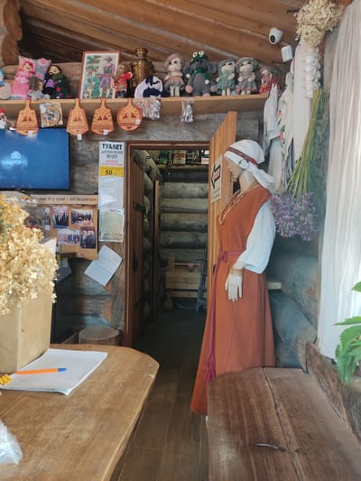 This image depicts the interior of a rustic building in Staraya Russa, Russia, characterized by log walls and wooden ceilings. The scene presents a reception or display area featuring cultural artifacts and a mannequin. In the foreground, a light brown wooden table is positioned to the left. On this table, a white spiral-bound notebook with an orange and blue pen resting on it is visible. A portion of a clear plastic bag and a brown cardboard container holding dried, light-colored floral material are also present on the table's left edge. The midground is dominated by a female mannequin standing on the right, facing towards the left. The mannequin wears a traditional costume consisting of a long-sleeved white blouse, an orange sleeveless tunic (sarafan) cinched with a red braided cord, a white head covering with a red and white patterned band, a multi-strand necklace of light brown disc-shaped beads, and a gold-colored cuff bracelet on its right wrist. To the left of the mannequin, a flat-screen television is mounted on a log wall, displaying a blue screen with white text. Above the television, a wooden shelf holds various handmade textile dolls and stuffed animals, including a pink doll, a green frog-like figure, and a grey doll. A framed picture depicting two individuals and a dog is also on the shelf, along with a brass samovar and several packaged items. Below the shelf, five wooden decorative plaques or cutting boards, some with animal motifs, are hung. Further left on the wall, below the television, a corkboard displays multiple photographs of men, some in suits, along with printed documents. Another white document is taped below these photos. Adjacent to the TV and to the left of a wooden doorway, a laminated sign in Cyrillic indicates "ТУАЛЕТ ДЛЯ ПОСЕТИТЕЛЕЙ МУЗЕЯ" (TOILET FOR MUSEUM VISITORS), stating "Бесплатно только во время посещения" (Free only during visitation) and "В остальных случаях 50 руб." (In other cases 50 rubles), with "50 руб." highlighted in yellow. The wooden doorway leads to an interior space with visible horizontal log walls and a wooden staircase descending into the background. To the right of the mannequin, bundles of dried plants and herbs hang from the wooden ceiling beams and log wall. These include white dried flowers (possibly hydrangeas), a string of white garlic bulbs, green reeds, and spherical clusters of dried purple flowers. A piece of white, possibly unbleached, fabric hangs vertically