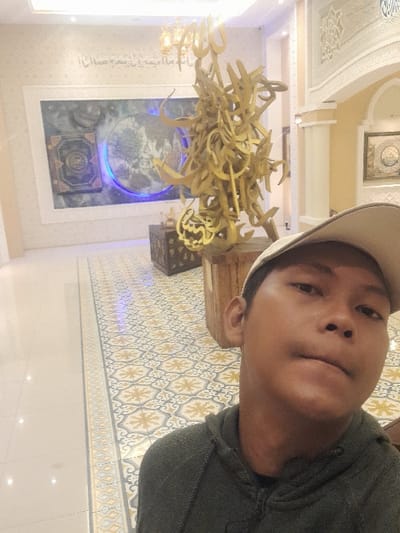 The image depicts an indoor scene in Glantengan, Indonesia. In the foreground, a young male individual, partially obscured, is shown from the chest up, wearing a light-colored baseball cap and a dark grey hooded sweatshirt, facing the camera as if taking a selfie. Behind him, centrally located, is a large, abstract golden sculpture composed of intertwined Arabic calligraphy, resting on a wooden plinth. Below the sculpture is an ornate, dark wooden chest with metallic accents. Above the sculpture, a golden chandelier with multiple light sources is suspended from the ceiling.
The background features a large rectangular wall art piece displaying a central circular motif with blue backlighting, resembling a globe or celestial body, alongside an open book or tablet on its left. Further Arabic calligraphy is visible on the wall above this artwork. The walls are light-colored and textured. The floor is covered with patterned tiles featuring blue, grey, gold, and white designs, leading to a lighter, plain tiled section on the left. On the right, ornate arched architectural elements are visible, suggesting a grand interior space, with another framed artwork partially visible in the distance. The scene is brightly lit by artificial light sources.