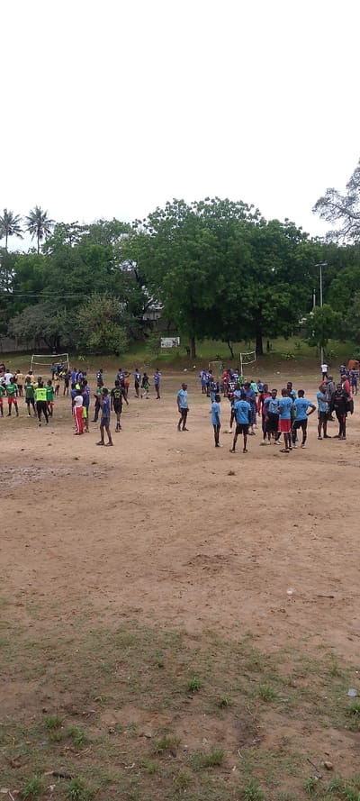 The image displays an unpaved outdoor sports field located in Tudor ward, Kenya, under an overcast sky. Approximately 50 to 60 individuals, primarily males, are scattered across the reddish-brown dirt field, which contains sparse patches of green grass and minimal debris, including a clear plastic bottle in the foreground.
In the background, a dense line of diverse green trees, including several palm trees on the left, forms a natural barrier separating the field from a visible white wall/fence and indistinct structures further behind. A tall light pole stands in the right background.
Two soccer goalposts are present on the field: a darker, smaller goal on the far left, and a larger white goal with a net positioned more centrally. A white net structure, consistent with a volleyball net, is also visible near the central goalpost.
The individuals are mostly dressed in athletic wear, including various colored jerseys and shorts. Notable groups include players wearing light blue jerseys with numbers (e.g., 15, 16, 36, 38) and others in lime green/yellow jerseys (one displaying "MAMY" on the back). Individuals are observed standing in clusters, walking, or in various states of readiness, with some appearing to be actively engaged in play or training near the left goalpost.