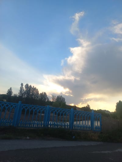The image captures a serene outdoor scene, likely during sunset, given the warm glow of the sun breaking through dramatic clouds. The sky is a dominant feature, transitioning from a clear, bright blue on the left to a mix of grey and golden-tinged clouds on the right. The sun's rays are visible, creating a luminous effect.
In the foreground, a bright blue metal fence with an intricate, decorative pattern runs horizontally across the frame, suggesting it might be a railing or barrier along a path or waterfront. Beyond the fence, a body of water is visible, reflecting the sky. The scene is bordered by lush greenery and trees, which are silhouetted against the bright sky, particularly on the right side of the image. A paved road or pathway runs alongside the fence on the far right, appearing somewhat wet, possibly indicating recent rain or dew.
There are no people or specific objects identifiable that indicate an activity or event. The overall mood is peaceful and tranquil, enhanced by the natural beauty of the sky and the surrounding landscape. The location cues of Kasansay, Uzbekistan, might suggest a rural or semi-rural setting with natural waterways and vegetation. No visible text is present in the image.