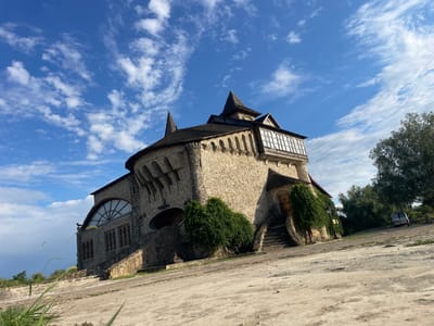 A low-angle, wide shot captures a peculiar, castle-like stone building under a bright blue sky with scattered white clouds. The building, with its multi-peaked dark roof and rough stone facade, is situated on a slightly elevated, uneven dirt and gravel lot. Green bushes and vines climb the sides of the structure, and a set of stone stairs leads to an entrance on the right. To the far right, partially obscured by trees, a white van is parked on a dirt road. The building appears to be located in a rural or semi-rural setting.