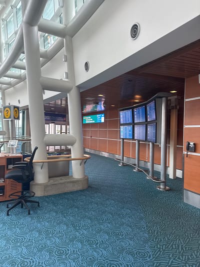This is an interior shot of an airport terminal. The scene is dominated by architectural elements like white pillars, a wooden paneled wall, and a patterned blue carpet. Several digital display screens show flight information, with some text visible indicating "Train to City" and "Sky Train Canada Line", suggesting a transit connection within the airport or to a nearby city. The lighting appears to be artificial, casting a neutral glow across the space.

There are no people visible in the image. The objects present include the display screens, a wall-mounted telephone, some seating (partially visible), and a rolling office chair. The overall impression is one of a modern, functional, and relatively empty public space, likely an airport or transportation hub. The time of day cannot be determined from the image. The location is identified as Richmond, Canada. The overall mood is calm and orderly, with no indication of any immediate activity or urgency.
