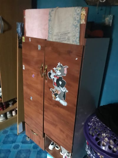 An interior scene in Oba-Ile, Nigeria, depicts a wooden two-door wardrobe positioned centrally. The wardrobe is constructed of a brown material, featuring golden-colored arched handles on its main doors and horizontal pull handles on two lower drawers. A large sticker depicting the cartoon characters Tom (in a tuxedo) and Jerry is affixed to the right main door, with several smaller, indistinct stickers present on both doors. The wardrobe appears to be mobile on small wheels. On its top surface, a folded pink fabric, a beige patterned fabric with text, and another patterned fabric are arranged. To the immediate left, a section of a light-brown wooden cabinet or shelving unit is visible, displaying multiple pairs of shoes on shelves. To the right of the wardrobe, a wall painted a solid blue is present, and a purple perforated plastic basket containing laundry or other items occupies the lower right foreground. Behind the wardrobe, a grey cabinet or partition is partially visible, with a red object observed on a surface above it. The floor is covered with blue and lighter blue patterned tiles. No individuals are present in the frame.