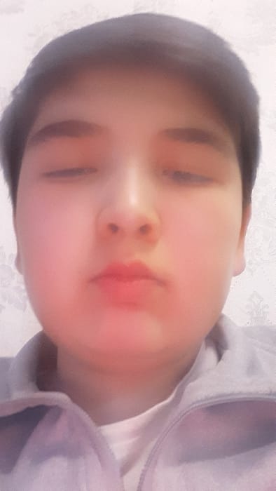 This image presents a close-up, head-and-shoulders portrait of a single young individual, taken indoors within the city of Pap, Uzbekistan. The subject has dark, short hair, and fair skin, with both eyes closed and lips visibly pursed. The individual is attired in a light-colored inner shirt beneath a light grey or lavender hooded jacket that is zipped up. The background is a light-toned wall featuring a subtle, repeating patterned wallpaper with floral or damask motifs.