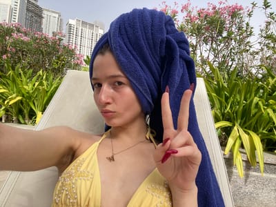 A woman wearing a yellow swimsuit with pearl embellishments and a gold necklace with a letter "M" pendant lies on a white mesh lounge chair with a dark blue towel wrapped around her head. She is making a peace sign with her left hand, showcasing long, red, pointed fingernails. The background includes plants and blooming pink flowers, and in the distance, high-rise buildings characteristic of the Business Bay area in the United Arab Emirates are visible.