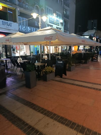 The image shows an outdoor seating area of a cafe at night, located on a pedestrian street in Niš, Serbia. The cafe has several tables and chairs set up under white umbrellas, some of which are branded with "Coca-Cola". A menu board with "Parcaffè" written on it is visible. In the background, buildings with illuminated signs and windows are seen. The street is paved with orange and dark-colored tiles, and streetlights illuminate the scene. There are no people visible in the immediate foreground or clearly engaged in any specific action. The overall atmosphere is quiet and appears to be late at night.
