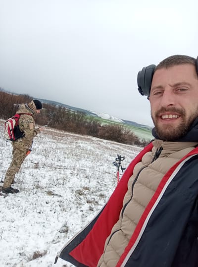 Two adult males are depicted in a snowy open field near the city of Drugan, Bulgaria, under an overcast sky. The individual in the foreground, on the right, is a male with a beard, wearing black over-ear headphones, a beige puffer jacket, and an open navy jacket with red lining. He is smiling and looking directly into the camera. In the mid-ground, on the left, a second male is visible, wearing a black beanie, a hooded camouflage suit, and a red and white backpack. He is holding a small electronic device, possibly a smartphone, in his hands, and a faint plume of vapor is visible near his face. A red/orange object is attached to his lower back. Between the two subjects, a piece of equipment with red poles, potentially a metal detector, is standing in the snow. The ground is covered by a thin layer of snow over sparse dry vegetation. The background features rolling hills with patches of green fields and barren bushes.