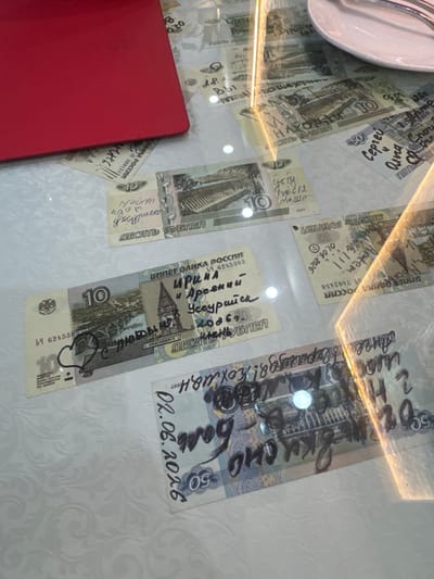 The image is an overhead shot of a glass table with several Russian banknotes laid out on it. Some of the banknotes have handwritten messages and drawings on them. The banknotes appear to be 10 and 50 ruble denominations. One banknote has the date "02.06.2026" written on it. The context of Suifenhe, China, is not evident from the visual information presented in the image. The scene is indoors, with a white textured tablecloth underneath the glass table. A red object, possibly a folder or book, is partially visible in the upper left corner. The lighting suggests a well-lit indoor environment.