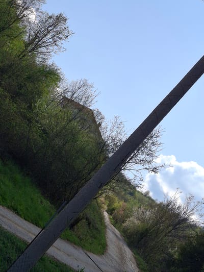 The image displays an outdoor, semi-rural landscape, consistent with the provided location context of Zverino, Bulgaria. The sky is bright blue with scattered white cumulus clouds.

A prominent, dark, and long rectangular object, likely a utility pole or a structural beam, extends diagonally across the frame from the upper right to the lower left. This object shows visible cracks or rough texture on its surface.

The majority of the scene is dominated by green foliage, including trees, bushes, and grass, which cover a sloping hillside. Some trees exhibit bare branches, suggesting early spring or late autumn, though vibrant green leaves are also abundant. Behind the denser vegetation on the left, a light-colored building, possibly an old stone or concrete structure with a sloped roof, is partially visible.

A dirt or gravel road/path winds its way uphill on the right side of the frame, disappearing into the distant trees and terrain. A section of what appears to be a wire fence is visible in the bottom right corner, intersecting the diagonal beam.

The clear lighting indicates it is daytime, likely mid-morning to late afternoon. There are no people or specific activities depicted, creating a peaceful and natural atmosphere. No visible text is present in the image.