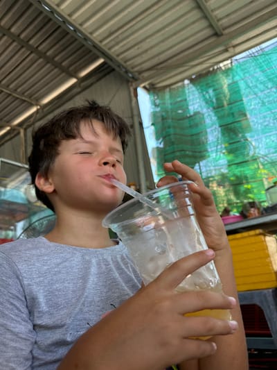 A young person is drinking from a clear plastic cup with a straw. The person is wearing a grey t-shirt. They are in an outdoor setting, possibly a market or roadside stall, with a corrugated metal roof overhead and green netting in the background. The lighting suggests it is daytime. The person's eyes are closed, and their face is tilted back as they drink, suggesting a moment of enjoyment or relaxation. Their hand is visible holding the cup. The overall scene is casual and appears to be a candid shot. There are some yellow crates visible to the left of the frame.