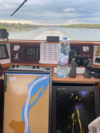 The image provides a detailed view from the navigation bridge of a boat, looking out over a wide river. The vessel appears to be actively underway, with instruments indicating navigation in progress during daytime under a partly cloudy sky. The location context of Ghindărești, Romania, places this scene on a major river, likely the Danube.
Through the window, calm river water stretches towards distant banks lined with trees. A wiper blade is visible in the lower-left corner of the exterior view.
Inside the helm, numerous objects and control systems are visible. A radar screen (Radio Holland) on the right displays yellow contacts and a blue track, indicating a range of "1.2/0.2km". Below it, a chart plotter running OpenCPN 5.6.2 shows a detailed map of a river with an "Island" and depth soundings like "sf dr 30.0" and "sf dr 34.0". The boat's current speed of "7.2 km/h" and a tracked path labeled "RUBISHIPS VI" are visible on this display.
On the wooden console, a pair of grey binoculars with a black strap is placed next to a plastic water bottle with a blue cap. The bottle's label reads "Балдаран ИЗВОРНА ВОДА 1.5Le" (Baldaran Source Water 1.5L). Various control panels and gauges are mounted on the console. These include a rudder position indicator (ST-4950) marked with "RUDDER POSITION", "PORT", and "STBD", as well as analog gauges for "V" (voltage) and RPM. A paper note lists engine RPMs from "500 RPM" to "1200 RPM" with corresponding fuel consumption figures ranging from "13L" to "250L". Other visible text includes "FA-150 PWR", "33014", "021049", "Radio Holland" on a control unit, and a sticker for "DSU DIVING ASSOCIATION NV UNDERWATER SURVEYORS".