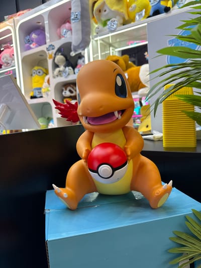 A close-up shot captures a Charmander figurine from the Pokémon franchise, seated on a light blue cardboard box. The figurine is predominantly orange with a yellow belly, large blue eyes, sharp teeth, and a distinctive flame-like appendage on its tail, which appears to be a stylized red wing in this instance. Charmander is holding a red and white Poké Ball in its hands, resting it on its lap.
In the background, a white shelving unit is filled with various plush toys and figurines, including what appear to be Minions, Hello Kitty, and other characters. The lighting suggests an indoor environment, possibly a retail store or an arcade. The scene does not provide any specific indicators of the city of El Paso, United States of America.