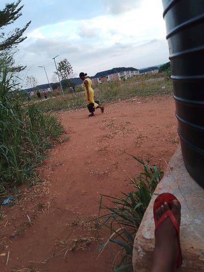 The image captures a scene in Ogboko, Nigeria. A woman wearing a yellow dress is walking on a red dirt path carrying a black bucket. In the background are buildings and street lights. In the foreground, the edge of a large water tank is visible, along with a foot in a red flip-flop. Grass and vegetation are also visible along the sides of the path.