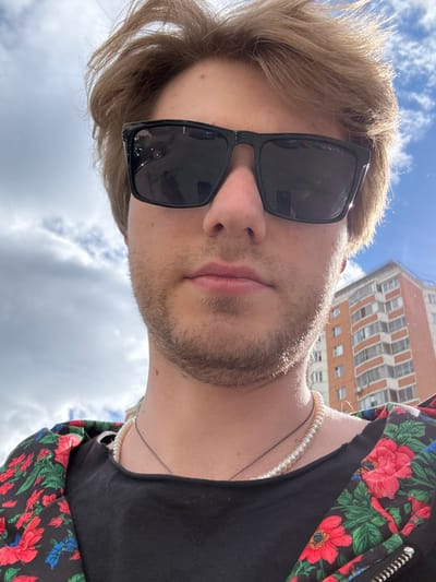 A medium-shot, street-level selfie is captured outdoors on a cloudy day. The subject, a young man with light brown hair and stubble, wears black sunglasses and a black t-shirt under a black jacket adorned with a vibrant floral print of red and blue flowers. He is also wearing a pearl necklace and a thin silver chain. The background features a tall, multi-story apartment building with orange and beige brickwork and visible windows. The sky above is a mix of clouds and patches of blue, with the sun casting a slight glare on the subject's hair and face. The image appears to have been taken in Mar'ino, Russia.