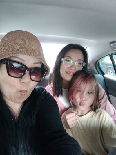 The image depicts three females in the backseat of a vehicle. The woman in the foreground, on the left, wears a straw hat, large sunglasses, and a black top with a pearl necklace. She is making a kissing face. To her right, a younger woman with glasses smiles, and next to her, a child with pink-streaked hair also makes a kissing face. The background shows the interior of a car, including the headliner and a portion of the rear seat. There are no discernible elements that indicate the location is Yahşihan, Turkey.