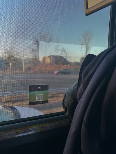 A bus window with a perforated sunshade features reflected buildings and sky. A dark draped garment obscures the top right corner. A green QR code sticker is adhered to the glass. An emergency exit instruction label is present with text in both English and Spanish, directing passengers to “Lift This Bar,” “Push Window Open.” The label also indicates the number “2” for the emergency exit procedure.