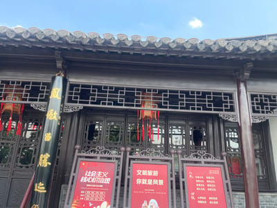 The image depicts a traditional Chinese building facade in the Hailing District of China. The building has a dark-colored wooden structure with intricate latticework, a tiled roof, and a hanging lantern. In front of the building are three red signs with white text, which contain written information in Chinese, likely about social values and tourism. A black pole with gold Chinese characters stands to the left of the signs. The sky is visible above the building, indicating it is an outdoor scene.