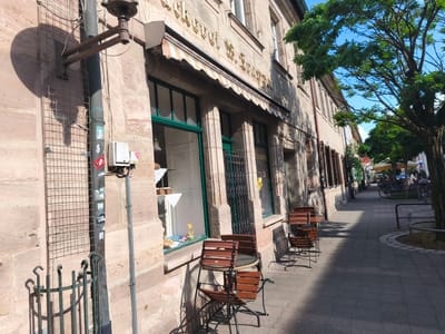 This image depicts the exterior of a bakery in Erlangen, Germany, as indicated by the text "Bäckerei W. Srappt" visible on the facade. The bakery has a traditional stone facade and a striped awning above its large windows. Several outdoor tables and chairs, made of dark wood and metal, are set up on the sidewalk in front of the establishment. The sidewalk is paved with cobblestones and is shaded by a large tree on the right side of the image. In the background, people are seated at tables outside another establishment, and a bicycle is parked nearby. The scene suggests a pleasant, sunny day in a European city, likely during daylight hours. The overall impression is one of a charming, active urban setting with a focus on outdoor dining and a local business.