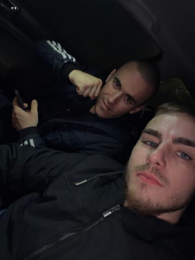 Two men are captured in a close-up indoor shot, likely within a vehicle due to the confined space and dark interior. The man on the right has fair skin, blue eyes, and a beard. He is wearing a black hooded jacket. The other man, positioned to the left and slightly behind, has a shaved head and is holding a dark-colored mobile phone. He is wearing a jacket with three white stripes on the sleeve. The lighting is dim, creating shadows and obscuring details in the background. There are no visible elements to indicate the city of Umarevtsi, Bulgaria.