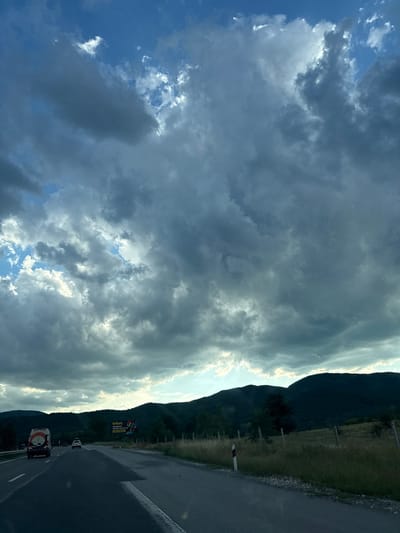The image depicts a view from a vehicle traveling on a paved road, likely a highway, heading towards a landscape with rolling hills and mountains in the background. The sky is dramatic, filled with dark, heavy clouds interspersed with patches of bright sunlight breaking through. The road itself has clear lane markings, and there are other vehicles on it, including a white van and a smaller white car further ahead. To the right of the road, there is a grassy verge with a wire fence and wooden posts. A large billboard advertising "efbet" is visible on the right side of the road, featuring colorful imagery and Bulgarian text. The overall impression is of a journey through a rural or semi-rural area under a dynamic, possibly approaching weather system. The location is identified as Gorno Varshilo, Bulgaria.