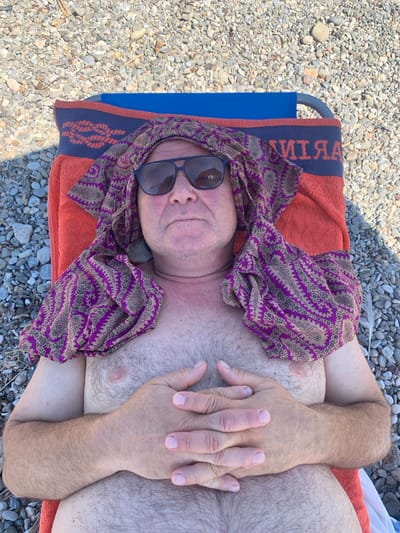 The image shows a person lying on a beach chair, covered by an orange towel and a patterned cloth. The person is wearing sunglasses and their arms are crossed over their chest. The scene is set on a beach with many small pebbles. The time of day appears to be sunny, as suggested by the sunglasses and the bright lighting. The location context is Pozzarello, Italy, which is consistent with a beach setting. No specific emotions are overtly displayed, beyond a relaxed pose. There is visible text on the blue part of the towel, which appears to spell "MARTINI" when read from right to left, with some letters obscured.