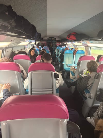 The scene depicts the interior of a passenger train car, observed from a seated position, looking forward down the central aisle. The train is potentially traveling through or located near Laucourt, France. The cabin features rows of passenger seats; those in the foreground have light gray frames with bright pink upper backrests, while seats further down the aisle are light blue with lighter green accents. Overhead luggage racks run along both sides, filled with various dark-colored bags and clothing items, including a distinct red backpack on the right side.

Multiple passengers are visible, primarily adults, seated and facing forward. In the left foreground, the arms and an open book held by a person with light skin are visible. Directly ahead, the back of a person wearing a white sweatshirt with partially visible text ("sion" and "HATTAN") and headphones is centered in the frame. Further down the left aisle, a woman with dark hair and glasses looks towards the right. On the right side of the train, a passenger with short, light-colored hair is seated, looking down at reading material, with a multi-colored bag positioned on a small table in front of them. The windows display a blurred green exterior, suggesting the train is in motion through a natural landscape. The overall environment is illuminated by natural light entering through the windows, creating a bright and active transit setting.