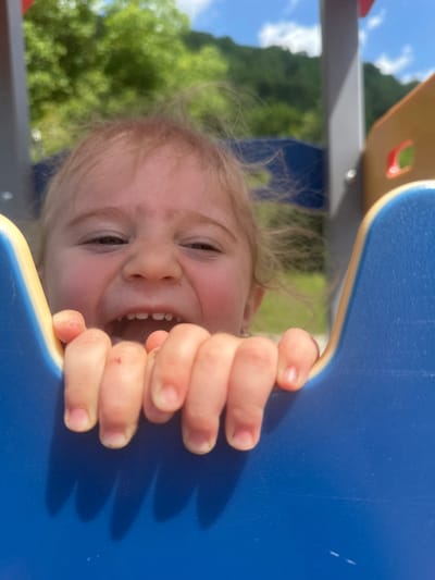 A close-up, low-angle shot captures a toddler with blonde hair leaning into a blue and yellow playground structure, their hands gripping the edge. The child's face is partially obscured but shows a wide, open-mouthed expression with visible teeth and rosy cheeks, eyes squinting. The background is blurred, revealing green foliage and a bright blue sky with white clouds, suggesting an outdoor setting on a sunny day. The specific location in Donchovci, Bulgaria, cannot be determined from the visual information provided.