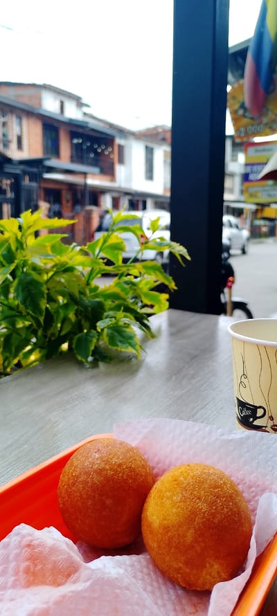 Two buñuelos, golden-brown fried dough balls, sit on a white paper towel in an orange plastic tray. A coffee cup with a coffee bean design is on the right. In the background, there's a green leafy plant and an out-of-focus street scene. Buildings and a black vertical column suggest an outdoor café or street food setting. The scene is brightly lit, giving the food a warm, inviting glow.