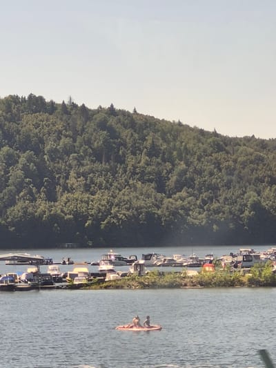 The image shows a body of water with boats docked on a small island or peninsula. The background is a densely forested hill under a clear sky. In the foreground, two people are sitting on a red raft in the water. The boats appear to be a mix of recreational vessels, with several parked along the shore. The environment suggests a summer day in a lakeside or riverside community. The location is identified as Pavlovka, Russia, which is a village in the Republic of Bashkortostan.