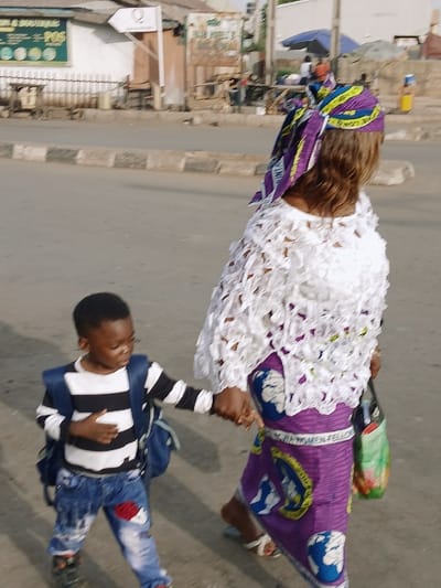A woman in a purple and white lace outfit and head wrap holds the hand of a young boy in a striped shirt and jeans. They walk along a street with buildings and signs in the background. The woman carries a green shopping bag. The boy looks up at her as they walk.