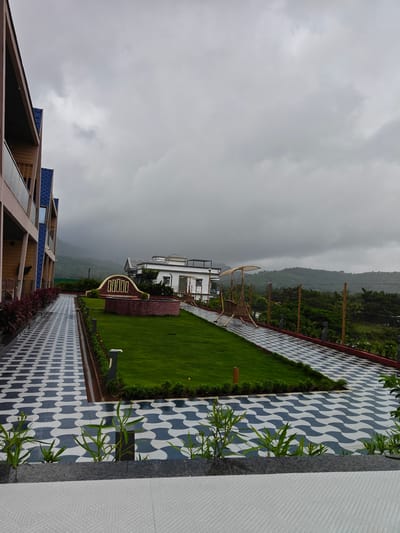 The image shows a resort in Ajivali, India, on a cloudy day. The resort features a modern building with a blue roof and balconies on the left side of the frame. A patterned walkway with blue and white tiles leads to a lush green lawn with a decorative brick structure in the center. Further back, a traditional swing set made of bamboo is visible, overlooking a landscape of green hills and trees under a cloudy sky. The overall scene depicts a serene and picturesque resort environment.