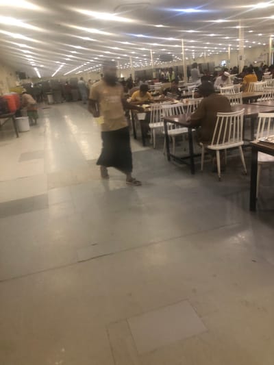 The image depicts a large, brightly illuminated indoor dining facility or cafeteria located in the city of النعيلة, Saudi Arabia. The ceiling features numerous evenly spaced, parallel rows of linear fluorescent or LED strip lights. The floor is covered with large, light-toned square and rectangular tiles, predominantly grey or off-white, with visible grout lines.
In the foreground, a male individual with a dark beard, wearing a light brown short-sleeved t-shirt with a discernible logo and a dark, long skirt-like lower garment (likely a lungi), is captured mid-stride, moving from left to right. He is wearing sandals and appears to be holding a light-colored cup or small plate in his right hand. His face and upper torso are blurred due to motion.
The midground and background are occupied by multiple rows of dark-surfaced rectangular tables, each paired with white chairs featuring vertical slat backs. Numerous adult males are seated at these tables, some engaged in eating or drinking from plates and cups. Their attire varies, including traditional thobes and head coverings, as well as casual shirts. In the far background, towards the left, industrial-style kitchen or serving equipment is visible, including large beverage dispensers and stainless steel counters, with additional individuals standing and moving in this area, suggesting a service or queuing point. The scene portrays a functional and busy communal dining environment.