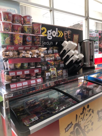 This is a vertical, eye-level, close-up shot of a display shelf in what appears to be a convenience store or small supermarket in Pilang, Indonesia. The primary subjects are rows of packaged food items, including cup noodles, snacks, and beverages, neatly arranged on shelves. The display is well-lit, suggesting it's daytime.

Prominently featured is a section dedicated to "2go! Seduh," which appears to be a brand of instant beverages or snacks, with some cup noodles such as "Kylafood Tagor Kuah CUP" and "Batagor Kuch Instan" also visible. Other brands like "Mie Sedap" and "Pop Mie" are also present, indicating a focus on quick meals. There are also various bottled drinks and some bagged snacks.

In the background, a stainless steel hot water dispenser with stacked paper cups suggests a self-serve beverage station. To the right, a glass-fronted refrigerator displays more products, possibly ice cream or cold drinks, with the word "BARU" (new) visible on a sign.

The scene is orderly and commercial, with prices displayed on some of the items, such as "Rp 8.000," "Rp 6.500," and "Rp 10.000." The overall impression is a well-stocked retail environment offering a variety of convenient food and drink options.
