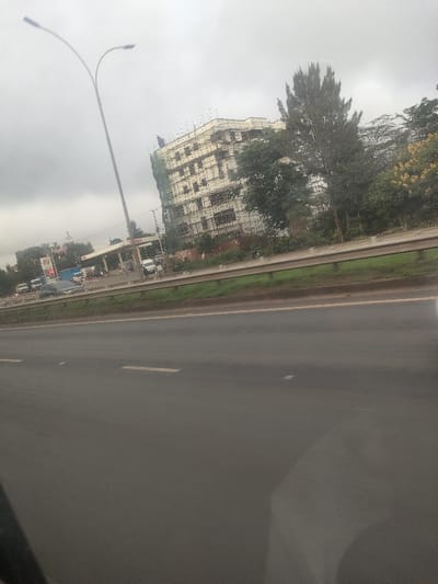 An exterior image captured from a moving vehicle displays a multi-lane asphalt road in the foreground, featuring white dashed lane markings. A grey metal guardrail separates the road from a grassy verge and a row of trees to the right. Beyond the trees, a multi-story building, approximately five to six floors tall, is under construction, evidenced by visible scaffolding on its facade and green safety netting on its left side. To the right of the building, a "TOTAL" branded gas station is visible, featuring a white canopy, several fuel pumps, and multiple parked vehicles, including a white van. A tall street light with two luminaires on a curved arm is positioned on the left side of the frame, extending towards the upper left. The sky is overcast and light grey, indicating cloudy conditions. This scene is located in Gatongora ward, Kenya. No specific individuals or distinct interactions are clearly discernible due to distance and image resolution.