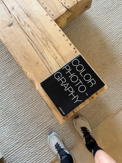 A high-angle, medium shot captures a book titled "COLOR PHOTOGRAPHY" placed on a textured, light-colored rug next to a rustic wooden beam. The book is black with white text. A person's legs and feet, wearing white athletic shoes and black socks, are visible in the lower right corner of the frame, indicating their presence near the objects. The scene is likely an interior setting with a focus on the natural textures of the wood and rug, and the graphic design of the book. The city of Artà, Spain is not depicted or implied in this image.