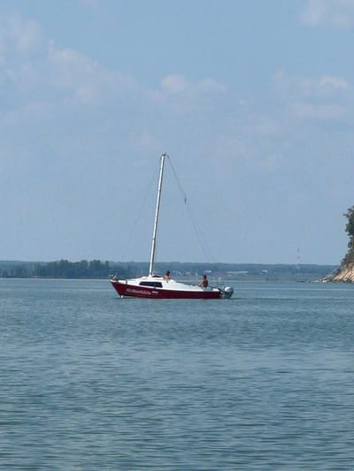 A red and white sailboat with two individuals aboard sails on a body of water. The boat is positioned in the center of the frame, its mast extending vertically towards a pale blue sky with scattered white clouds. The water's surface exhibits ripples, indicating a light breeze. In the background, a tree-lined shore is visible, with a distant structure resembling a radio tower on the right. The scene appears to be set in Bystrovka, Russia. The boat's hull displays the inscription "AB2B44RUS54". One person is visible sitting on the deck, while another stands near the stern.