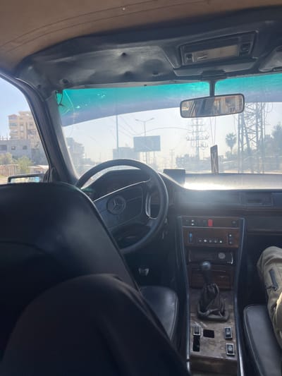 The image displays the interior of an older model car, likely a Mercedes-Benz, viewed from the rear passenger side, looking forward towards the dashboard and windshield. The image is oriented vertically, showing the car's left side at the top and its right side at the bottom, but the description will assume a standard horizontal orientation for clarity.

The primary focus is the car's cockpit. A black steering wheel with a visible Mercedes-Benz logo is prominent on the left side, indicating the driver's position. The dashboard is dark-colored, and the center console features a manual gear shifter with a black leather or vinyl boot. Surrounding the gear shifter and extending across the dashboard are panels with a worn wooden trim. These panels contain various buttons and controls, some of which appear to be for climate control or the audio system, while others are blank or show signs of wear.

In the foreground, a person's leg and lap are visible, clad in dark trousers, suggesting a passenger in the rear seat. Further into the car, another person's leg, wearing khaki-colored pants, can be seen on the front passenger seat. The car's seats are black, likely leather or vinyl. The headliner on the passenger side (viewer's right) is a light tan color.

Through the front windshield, a bright, sunny daytime scene is visible. The sky is clear, and strong sunlight streams into the car, creating bright highlights on the dashboard and windshield. The windshield itself shows significant damage, with multiple cracks spreading across the glass, particularly noticeable in the lower-left corner (from the driver's perspective). The top portion of the windshield appears to have a greenish-blue tint.

The exterior view reveals an urban or semi-urban environment. In the distance, several buildings are visible, including one that appears to be under construction or has exposed concrete. Power lines, large metal structures resembling electricity pylons or parts of a bridge, and streetlights are scattered across the horizon. A lone palm tree stands out against the sky. A rearview mirror is mounted on the windshield, reflecting some of the interior. The A-pillar on the driver's side (left of the windshield) is dark and shows some signs of wear.