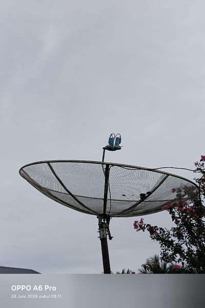 The media file shows a satellite dish against a cloudy sky. The dish is large and metallic, with a mesh surface supported by a dark metal pole. Attached to the dish are two blue components, likely part of the receiver. To the right of the dish, a branch of a tree with small pink flowers and green leaves extends into the frame. In the background, a roof of a building is partially visible on the left. The sky is overcast and appears to be a pale grey. The lower part of the image contains text that reads "OPPO A6 Pro" and "26 Juni 2026 pukul 08.11", indicating the device used to capture the photo and the date and time it was taken. The overall scene suggests an outdoor setting, possibly a residential area in Rheum Barat, Indonesia, on a cloudy morning. There are no people visible in the image.